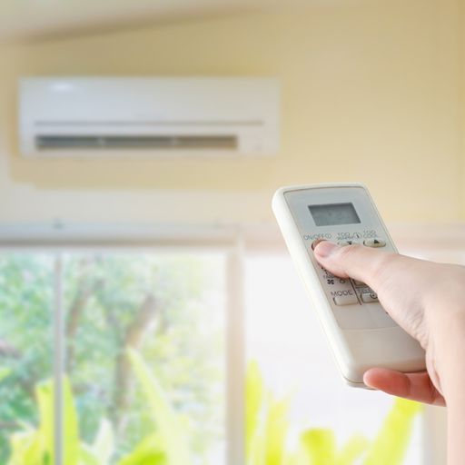 Image is a photograph from the POV of a person holding the remote for their ductless minisplit. In the bottom right corner is their hang holding the remote and pointing it at their ductless unit mounted on a yellow wall above a window.