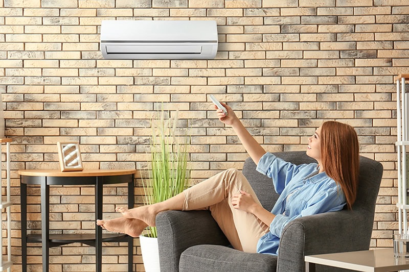 How Much Does it Cost to Install a Ductless Mini Split? Young woman switching on air conditioner while sitting in armchair at home.