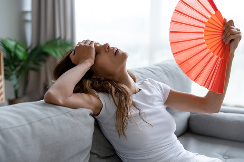Why Is My AC Blowing Hot Air? Photo of a young woman fanning herself off to cool down on her couch.