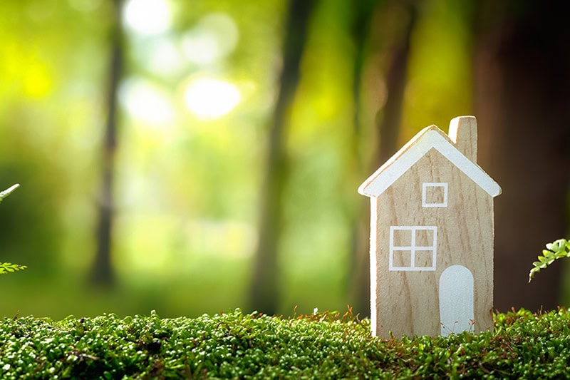 What Does a Geothermal System Cost? Photo of a wooden toy house on grass with a green forest background.